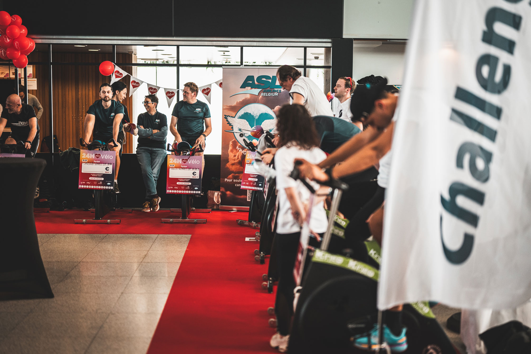 Vue d'un événement caritatif "24h Vélo" de Télévie à l'aéroport de Liège (LGG). Plusieurs participants sont installés sur des vélos d'appartement alignés sur un tapis rouge. En arrière-plan, on aperçoit des ballons rouges, des banderoles et une affiche de la compagnie ASL Airlines Belgium.